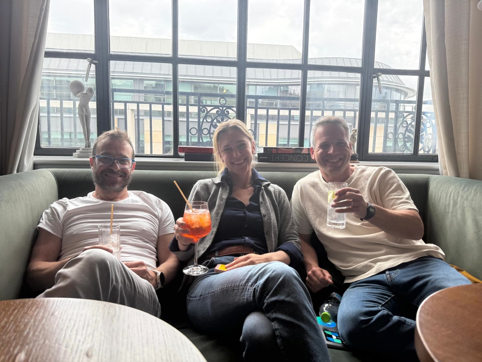Three people relaxing with cocktails on a sofa by tall windows overlooking a rooftop