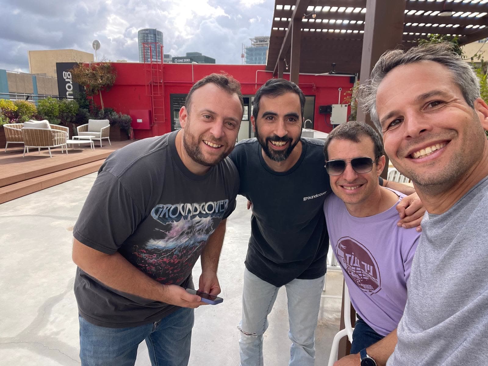 Four men posing arm in arm on a sunlit rooftop terrace with the city behind them
