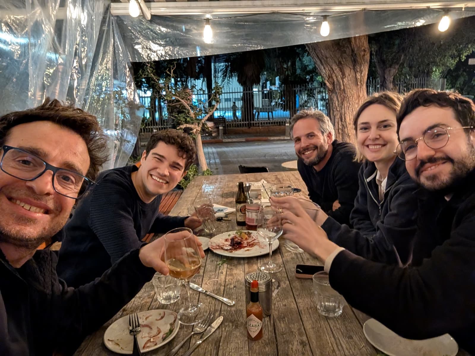 Five colleagues raising drinks around a wooden table on a covered outdoor terrace at night