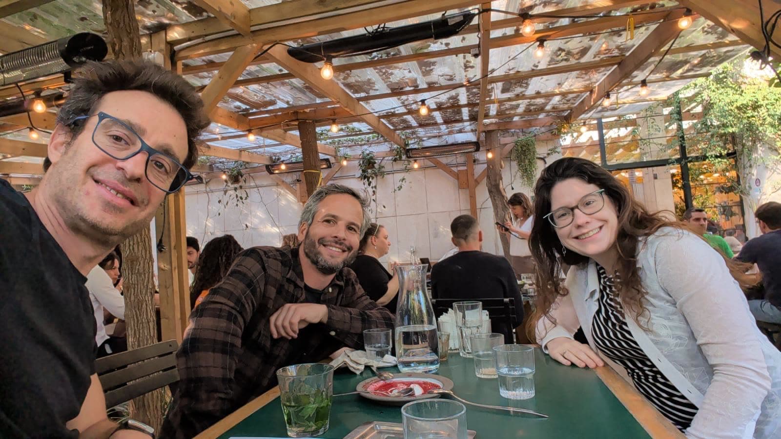 Three people at a long table under a pergola strung with lights at a busy restaurant