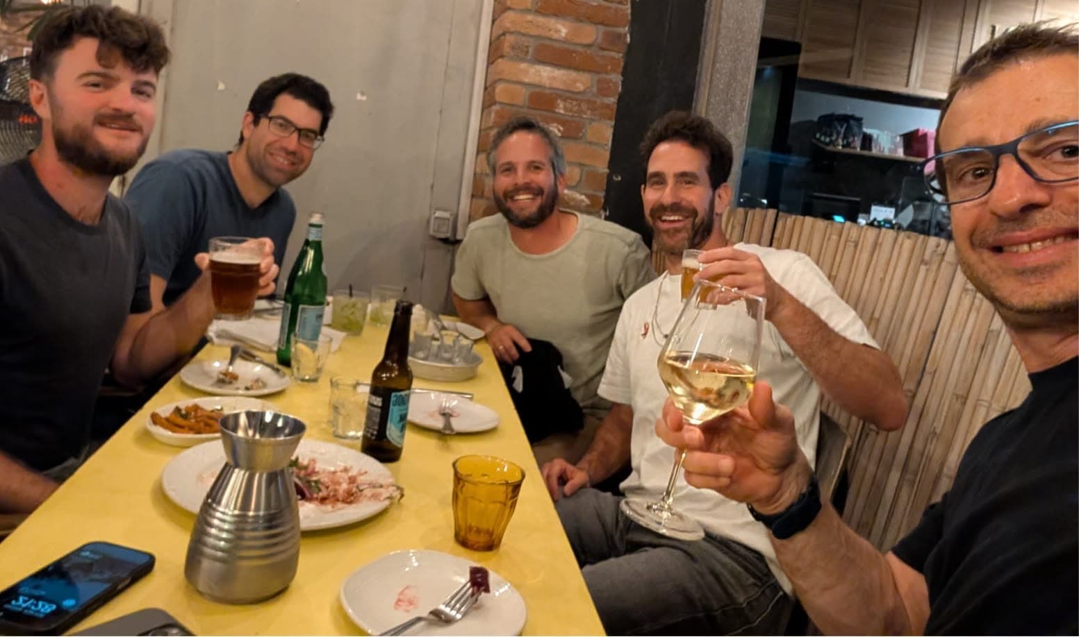 Five people at a long yellow table with beer and wine, toasting toward the camera
