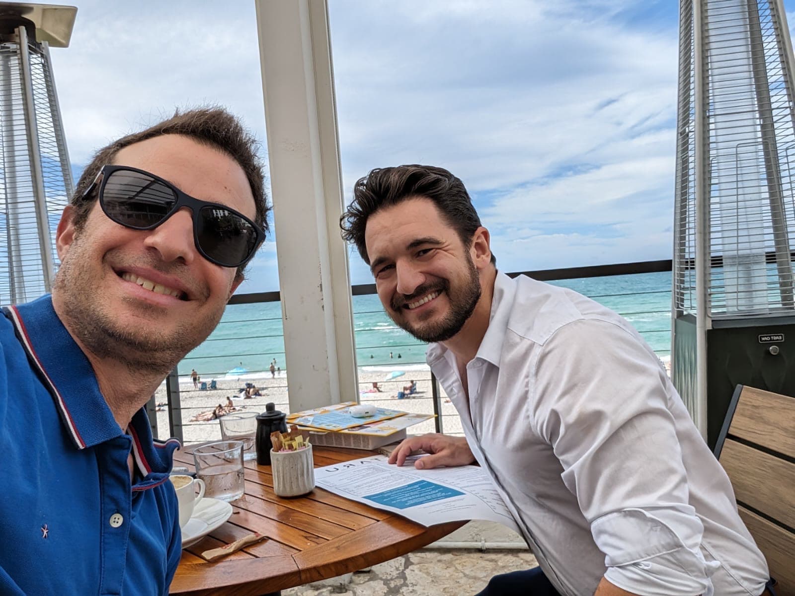 Two men at a seafront table with papers spread out, the beach and sea behind them