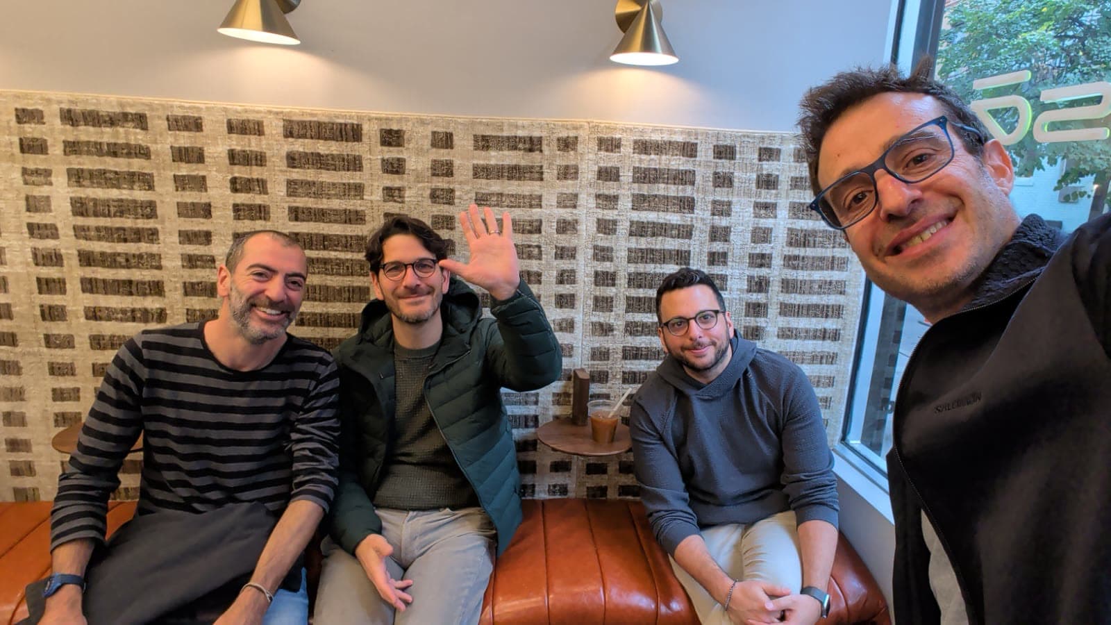 Four men gathered on a tan leather sofa in a cafe, one waving at the camera